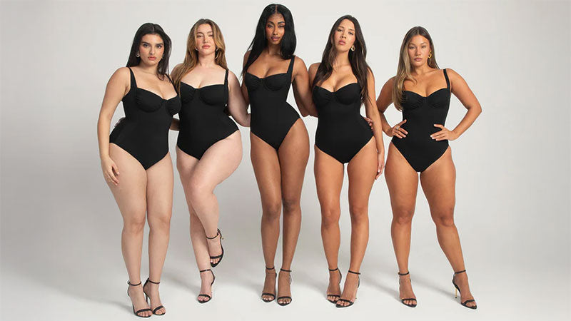 Five women of diverse body types stand confidently in black shapewear one-piece swimsuits against a plain background, conveying empowerment and inclusivity.